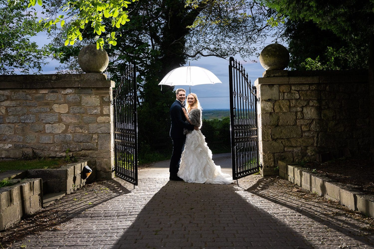 A bride and groom stand under a lit umbrella in the darkness, by a gate at the end of a path - taken by Hertfordshire Wedding Photographer Tim Payne