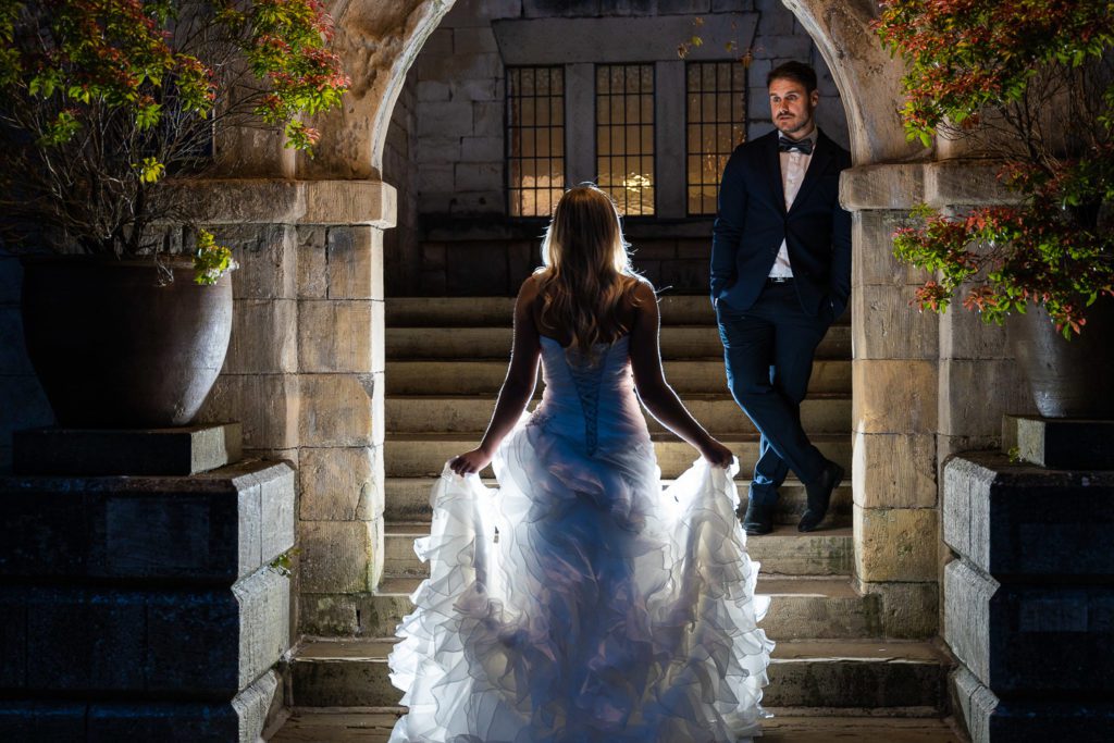 why pay for a professional wedding photographer - a bride is approaching the steps to a manner house, her back to the camera, backlit, her groom stands below an archway at the top of the steps