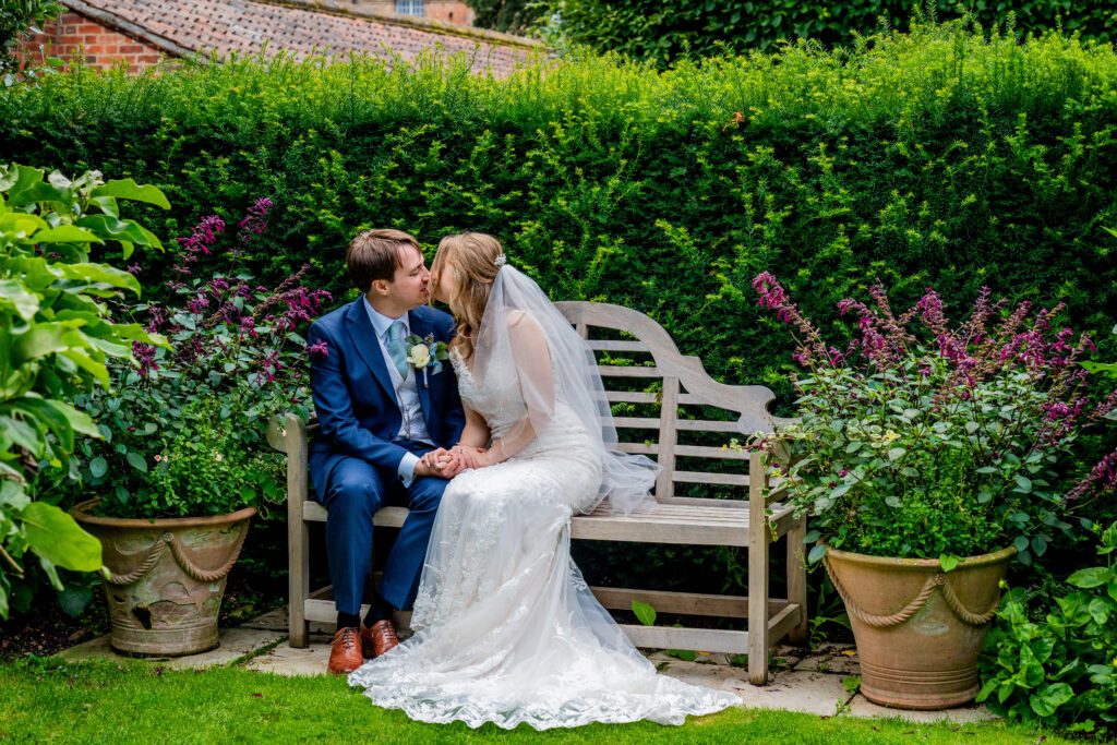 why pay for a professional wedding photographer - a bridal couple sit on a wooden bench in a formal garden and steal a kiss