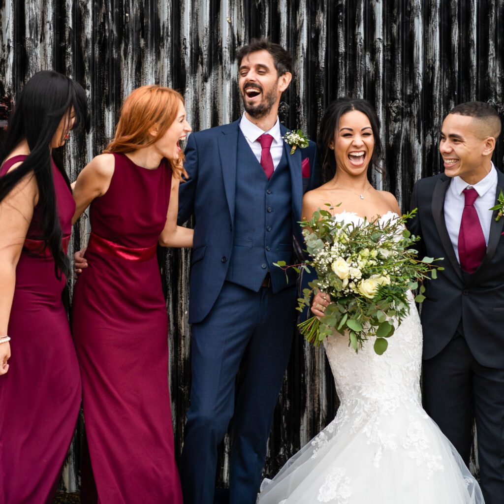 why pay for a professional wedding photographer? - image of a bride and grrom with bridesmaids in claret red and a best man all laughing.