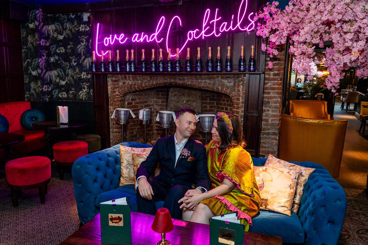 award-winning wedding photographer - a bridal couple, the groom in a navy suit, the bride in a short bright gold dress, sit on a blue sofa in a bar surrounded by pink blossoms on the ceiling and a neon sign above the fireplace stating love and marriage.