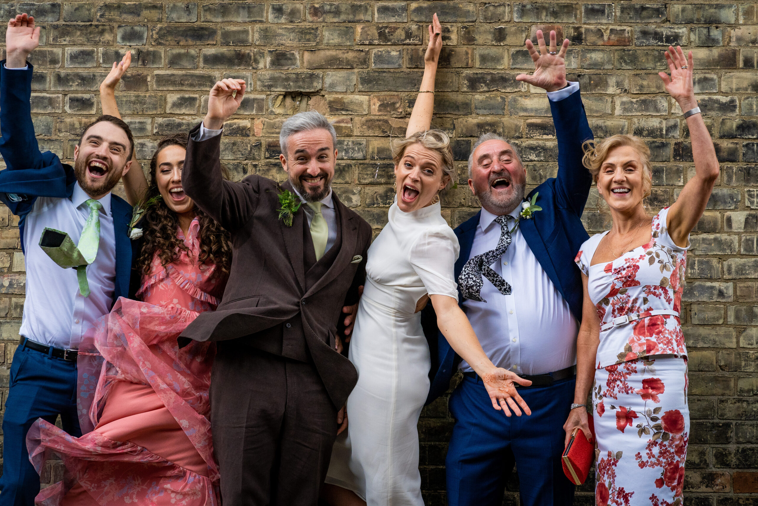 A bridal party celebrating with their arms up in the air and joyful faces against a brick wall