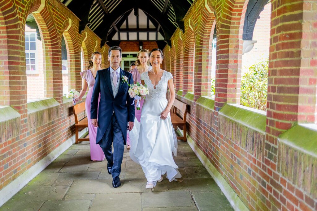 Bride walking through the cloisters to her church with her father. Tim Payne Photography a Hertford wedding photographer.