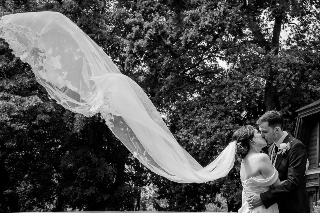 Black and white image of a bride and groom having a kiss at Fanhams hall, Ware with her veil blowing in the wind.Tim Payne Photography a Hertford wedding photographer