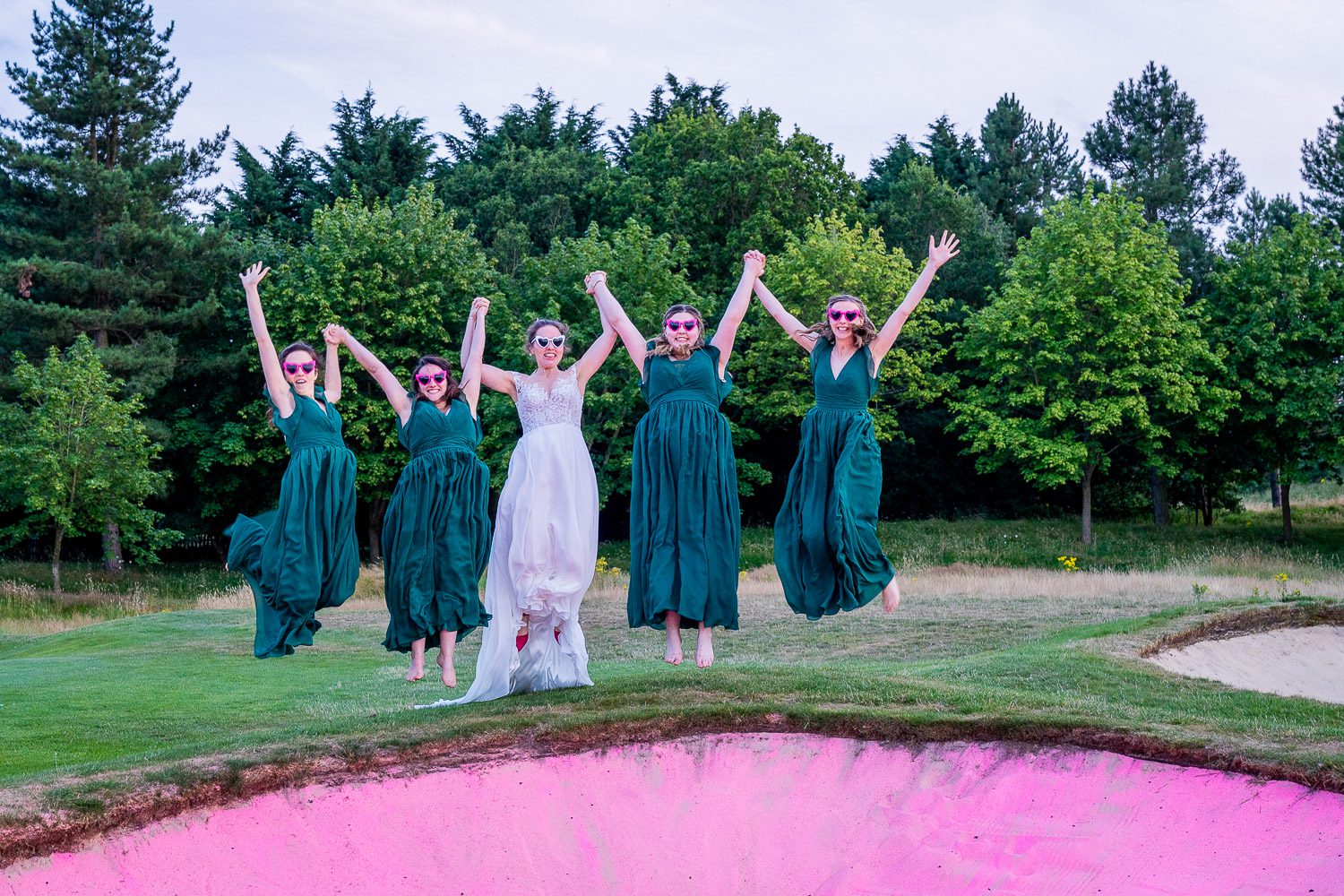 Bride and her bridesmaids jumping on the top of a sand bunker with pink sand, at Essendon country club. Tim Payne photography, a Hertfordshire based wedding photographe