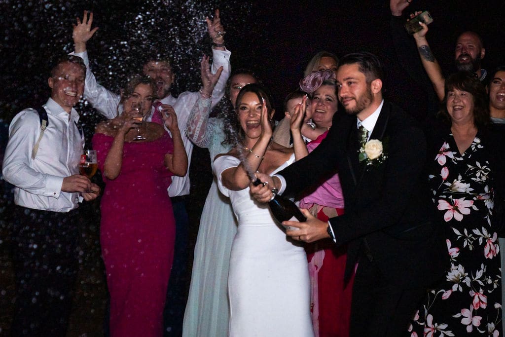 A groom, next to his bride is spraying champagne in the dark, their guests are cheering behind them