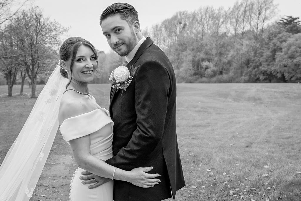Black and white of bride and groom holding each other, looking to camera with the golf course behind them