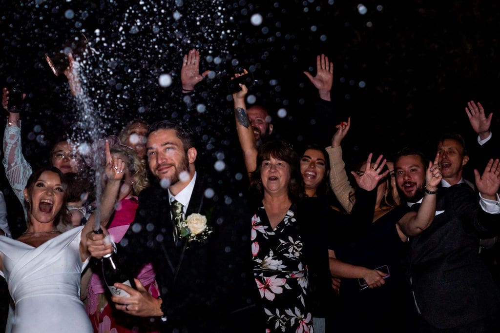 A groom sprays his bride and guests with champagne in the night time, everyone is cheering
