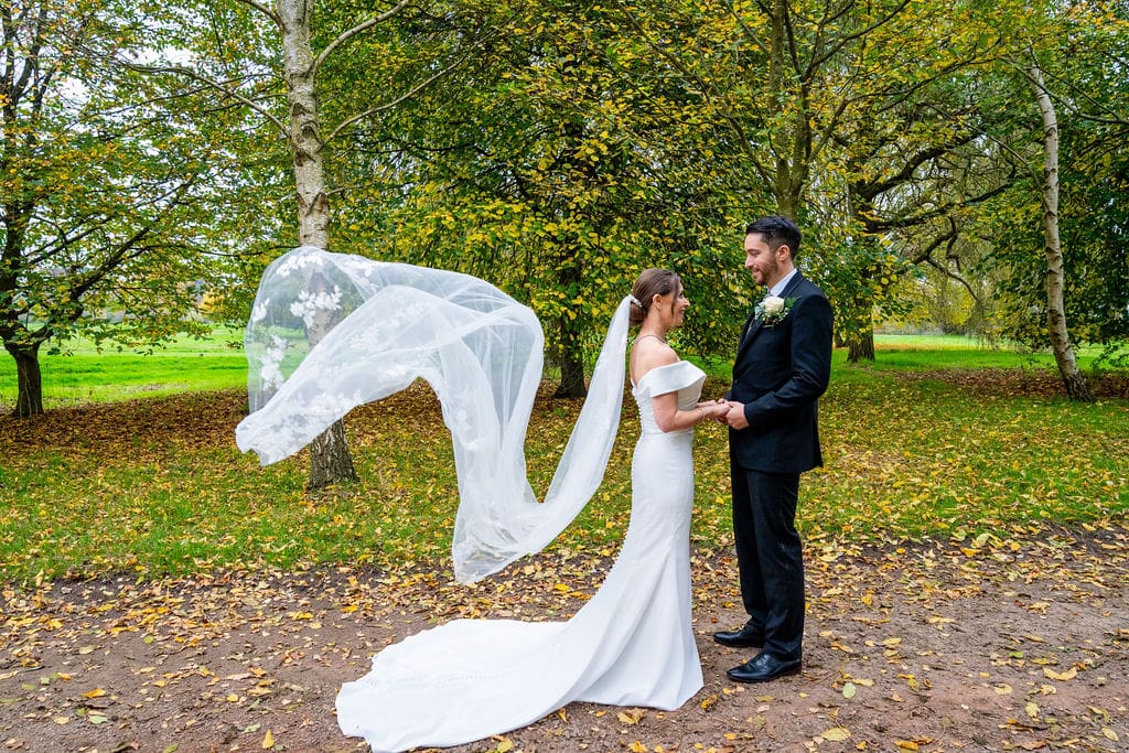 Bride and groom in a woodland setting, facing each other, holding hands, the veil wafting in the breeze