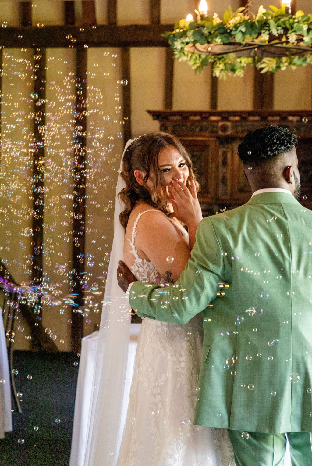 A bride turning back to look at her guests during the ceremony, laughing as bubbles float in the air around her, her groom in a green suit has his arm around her waist and his back to the camera