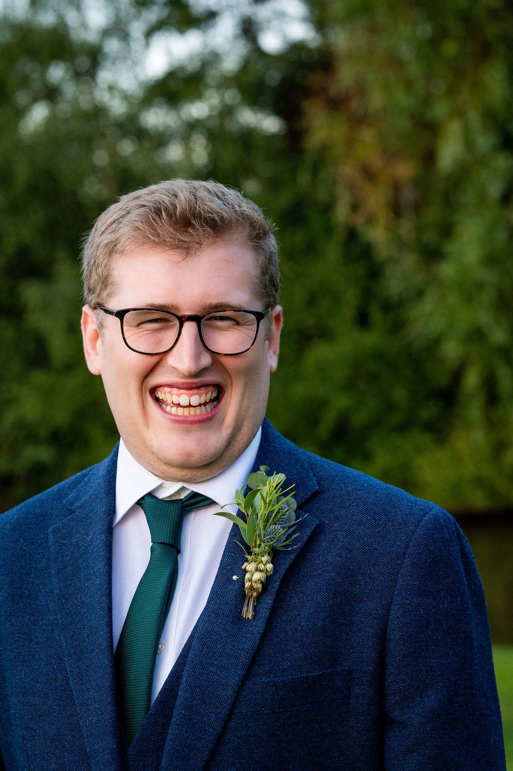 Close up portrait of a groom wearing glasses and a blue suit smiling at the camera