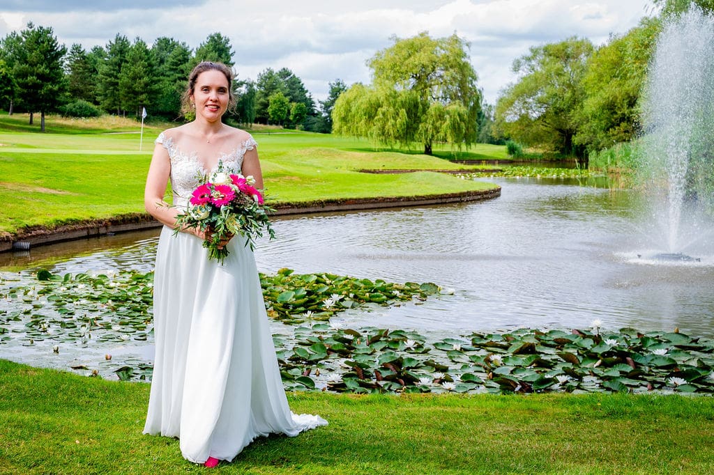 Traditional dressed bride with pink floral bouquet is standing in front of a lake with water lillies and a fountain at Essendon Country Club wedding venue