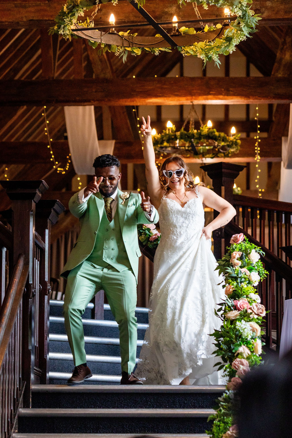 Bride and groom wearing sunglasses, dancing down a dark staircase with oak beams on the walls behind them