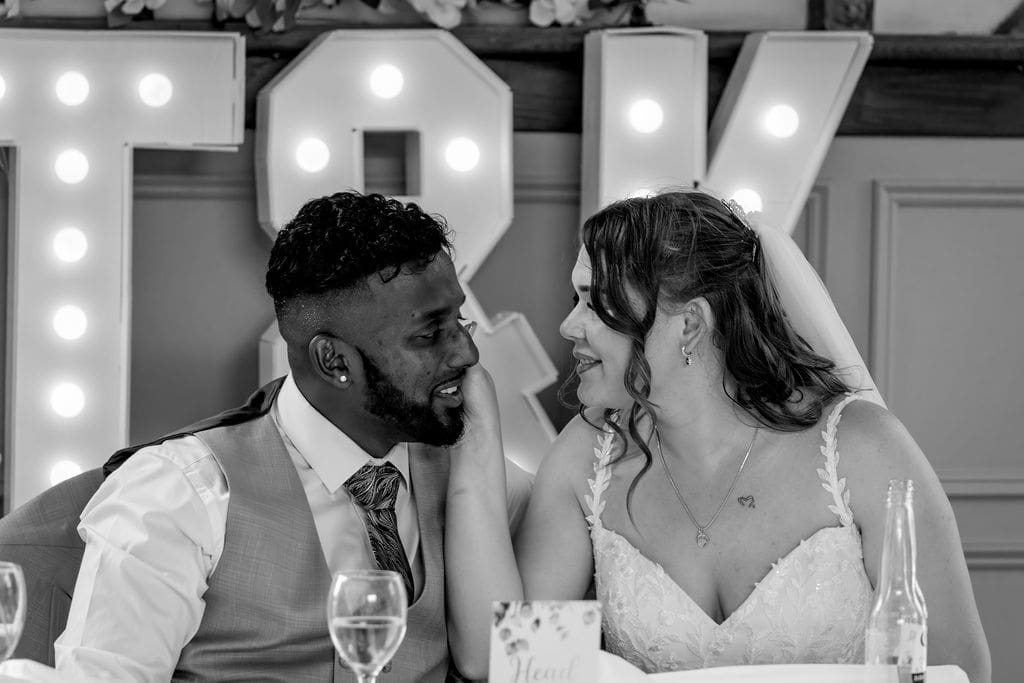 Black and white of a bride and groom sat at the table, she is touching his face gently as they gaze at each other, large lit up letters T&K are behind them