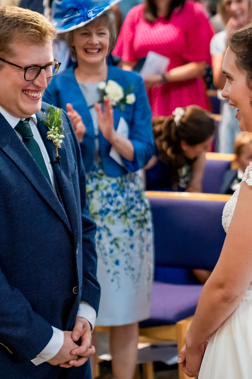 Bride and groom smiling at each other after saying their vows in church, groom's mum in a blue outfit is to the left of the groom's shoulder