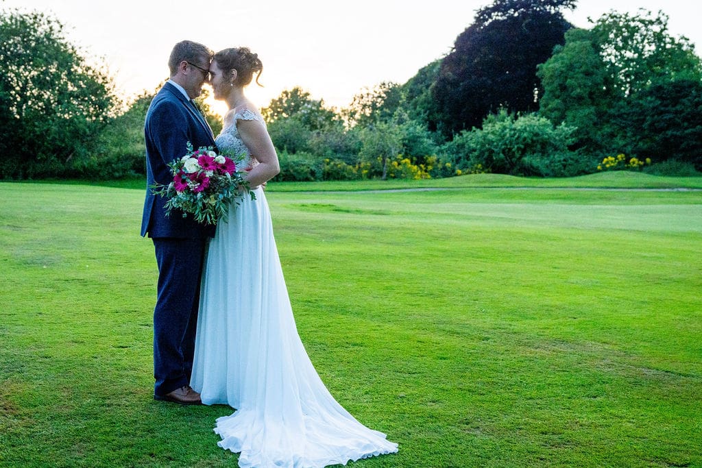 A traditional bride and her groom in a 3-piece blue suit hold each other with foreheads touching, they are on a green with trees and shrubs in the background