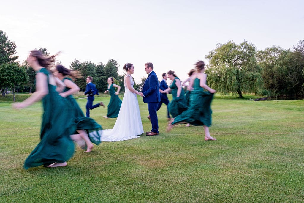 Bride and groom in the centre on a green, with blurred images of their wedding party running past
