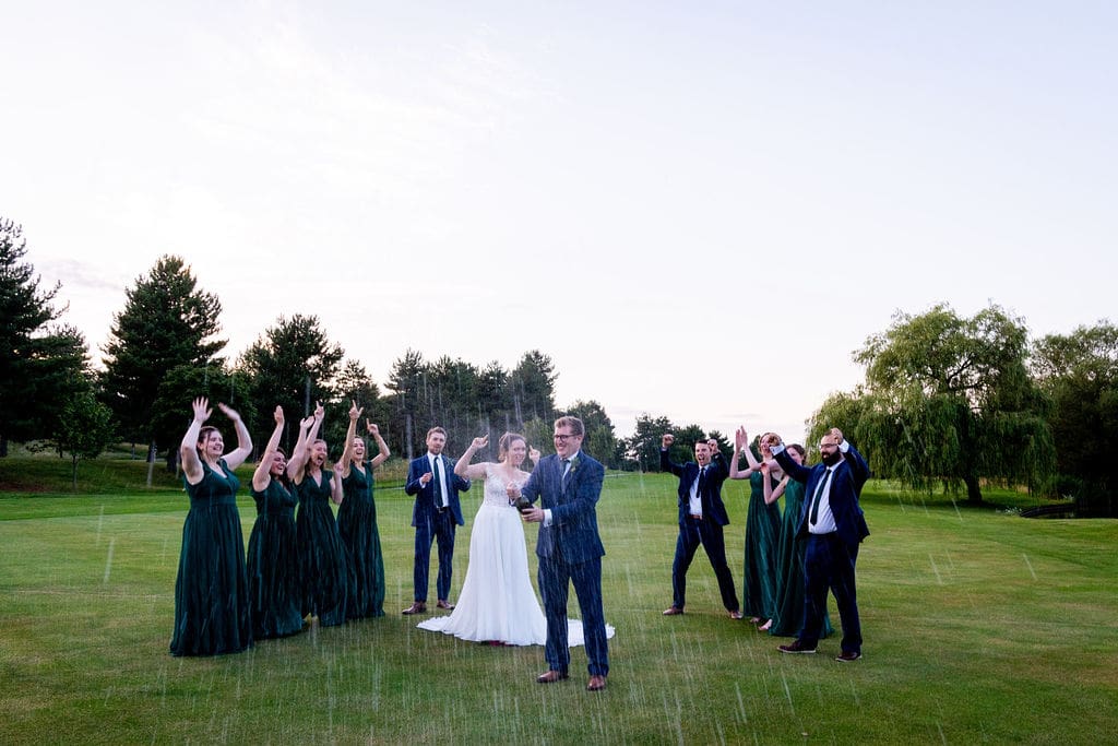 A groom sprays champagne with his bride by their side and surrounded by their groomsmen and bridesmaids