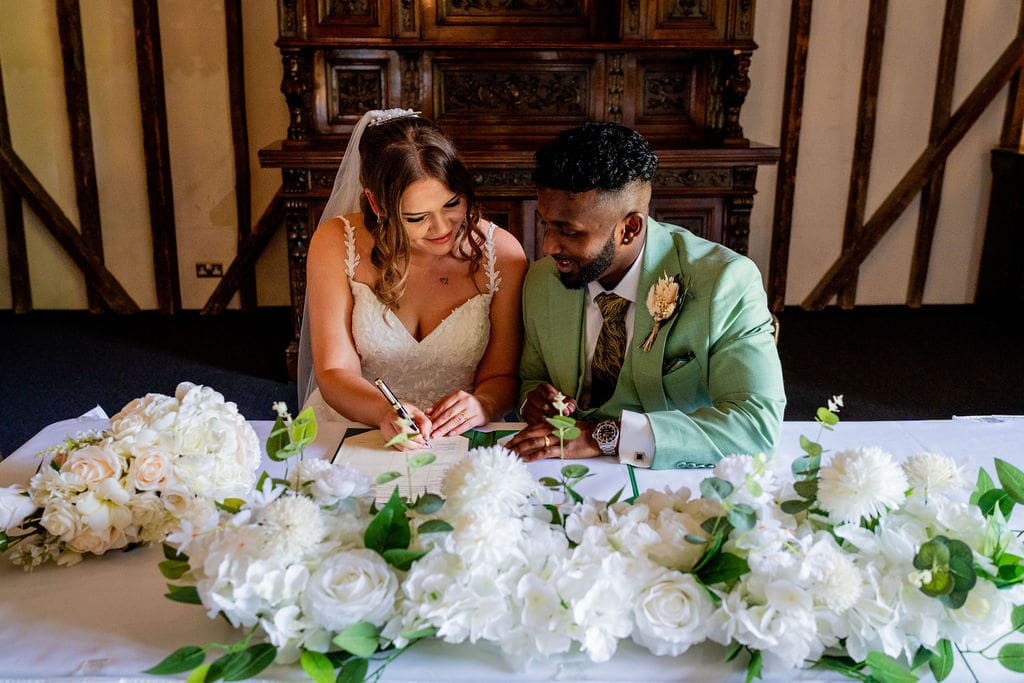 Traditionally dressed bride and a groom in a light green suit sit at a table with white flowers on to sign the register