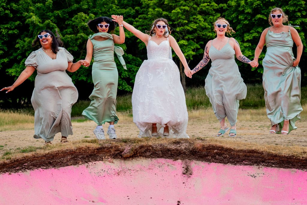 A bride, with 4 bridesmaids (2 either side) wearing sage green dresses and all wearing sunglasses are jumping into a pink bunker at Essendon County Club wedding venue