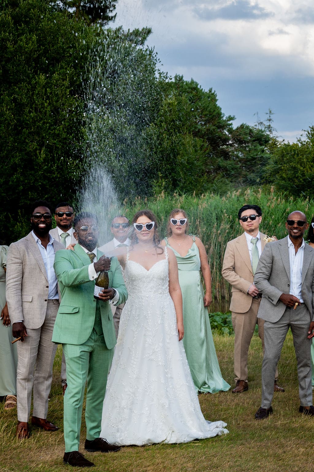 A groom in a light green suit is spraying champagne, with his traditionally dressed bride at his side and the wedding party behind them