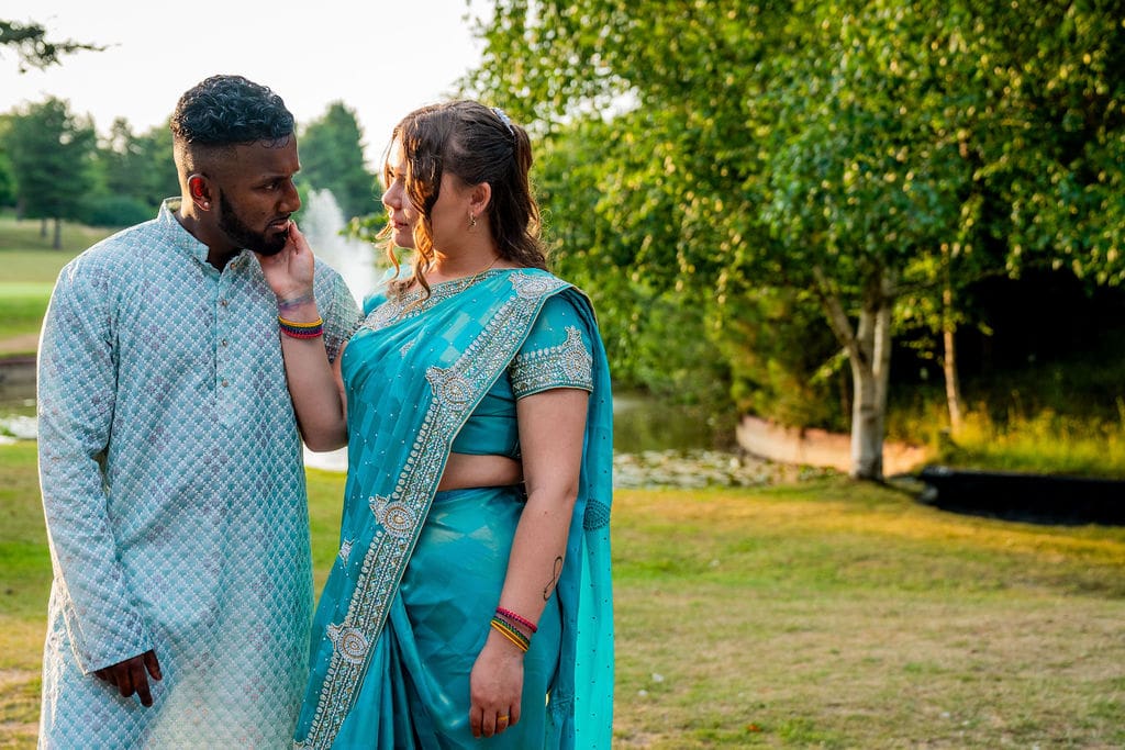 A bride and groom dressed in traditional blue Indian outfits look into each other's eyes with a lake, fountain and trees behind them