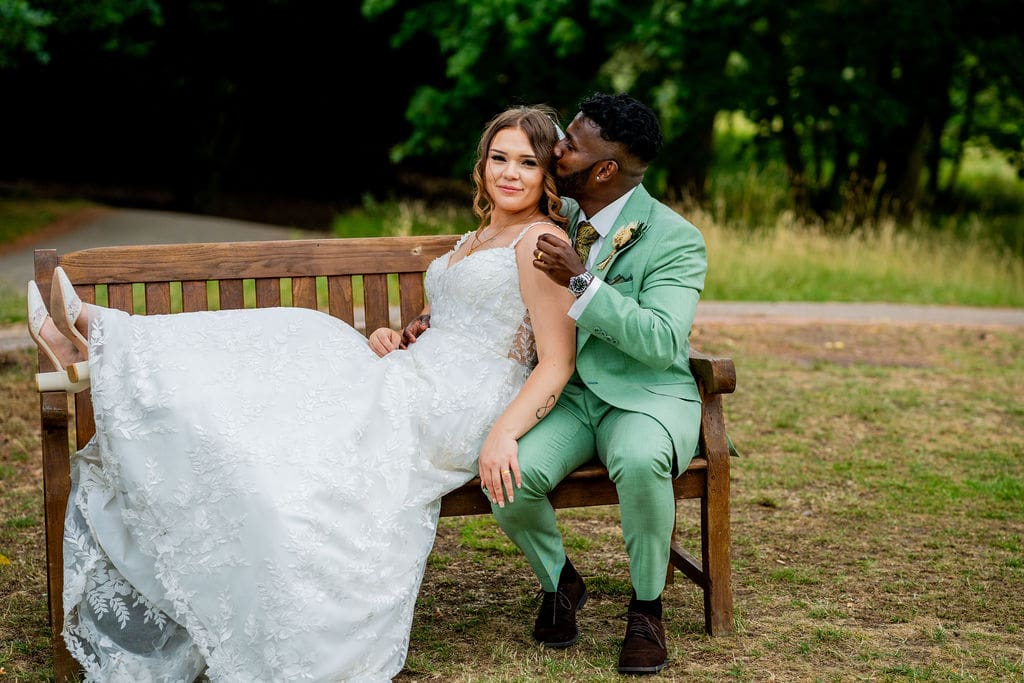 A traditional bride stretches out on a bench with her feet up, leaning against her groom, dressed in a light green suit.