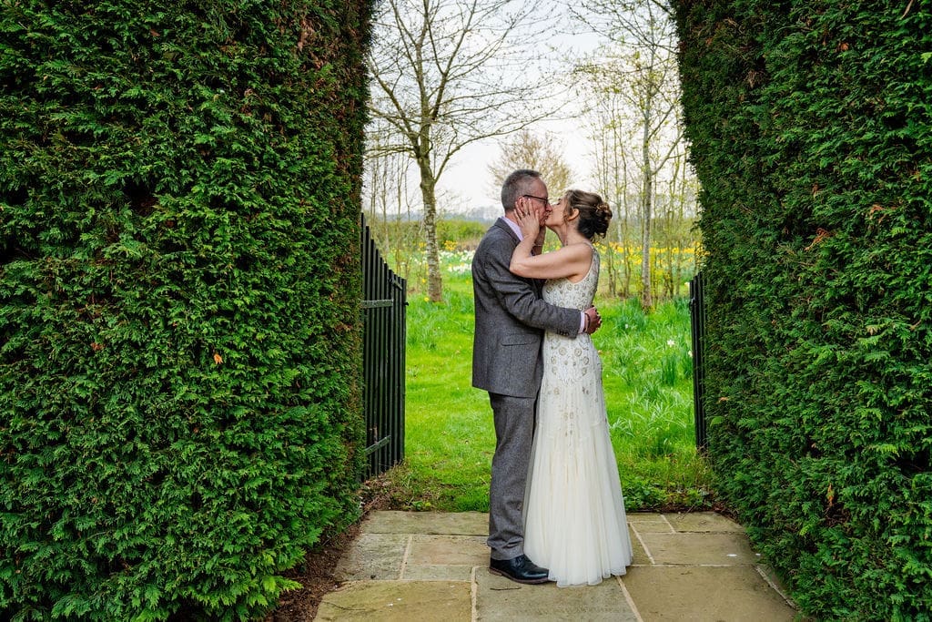 Wedding day photography - a bride and groom kiss within the arch of a large hedge